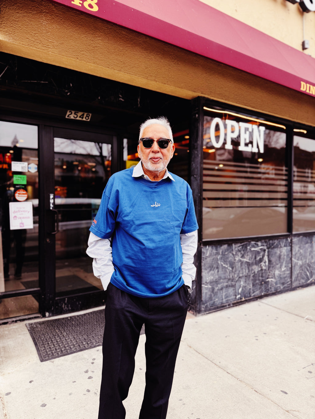 Man in a blue sweatshirt standing in front of a store with an 'Open' sign.